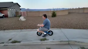 adult man sitting on a very small children's bike on a sidewalk in front of a block wall