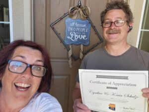Cindy and Kevin Schulze smile in front of their home while holding a certificate of appreciation from the Arizona Department of Child Safety recognizing 15 years of service to children and families.
