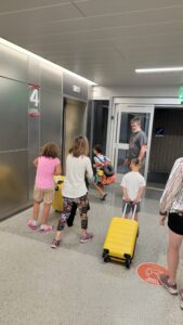 A family moves through an airport with bright yellow luggage while a man waits near the elevator doors and children walk ahead carrying travel bags.