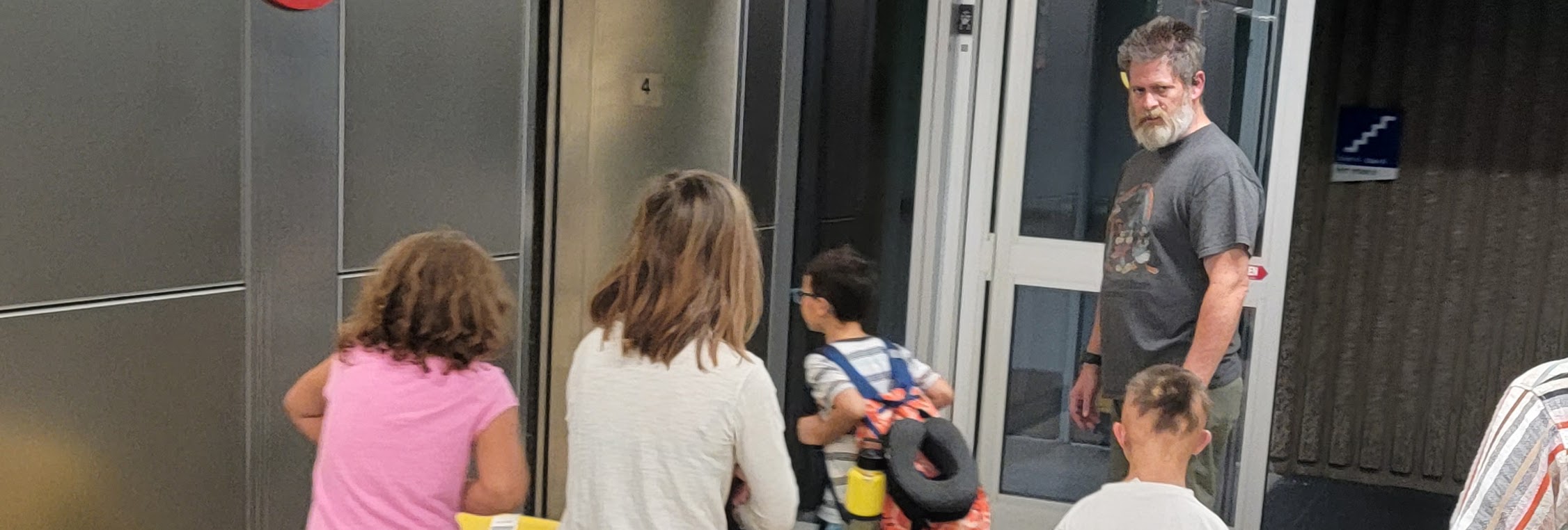 A family moves through an airport with bright yellow luggage while a man waits near the elevator doors and children walk ahead carrying travel bags.