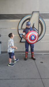 Ben stands talking with Captain America in front of a large Avengers symbol, looking up at him during a character interaction.