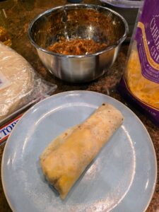 Breakfast burrito on a light blue plate beside tortillas, shredded cheese, and a bowl of filling on a kitchen counter.
