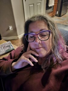 A close-up photo of a woman resting with her eyes closed, wearing purple-and-black glasses and a burgundy sweatshirt, her hand gently against her face. She appears quiet and tired, sitting indoors in a home setting.