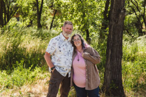 Cindy and Kevin stand close together in a sunlit wooded area, smiling at the camera with trees and tall grass surrounding them.