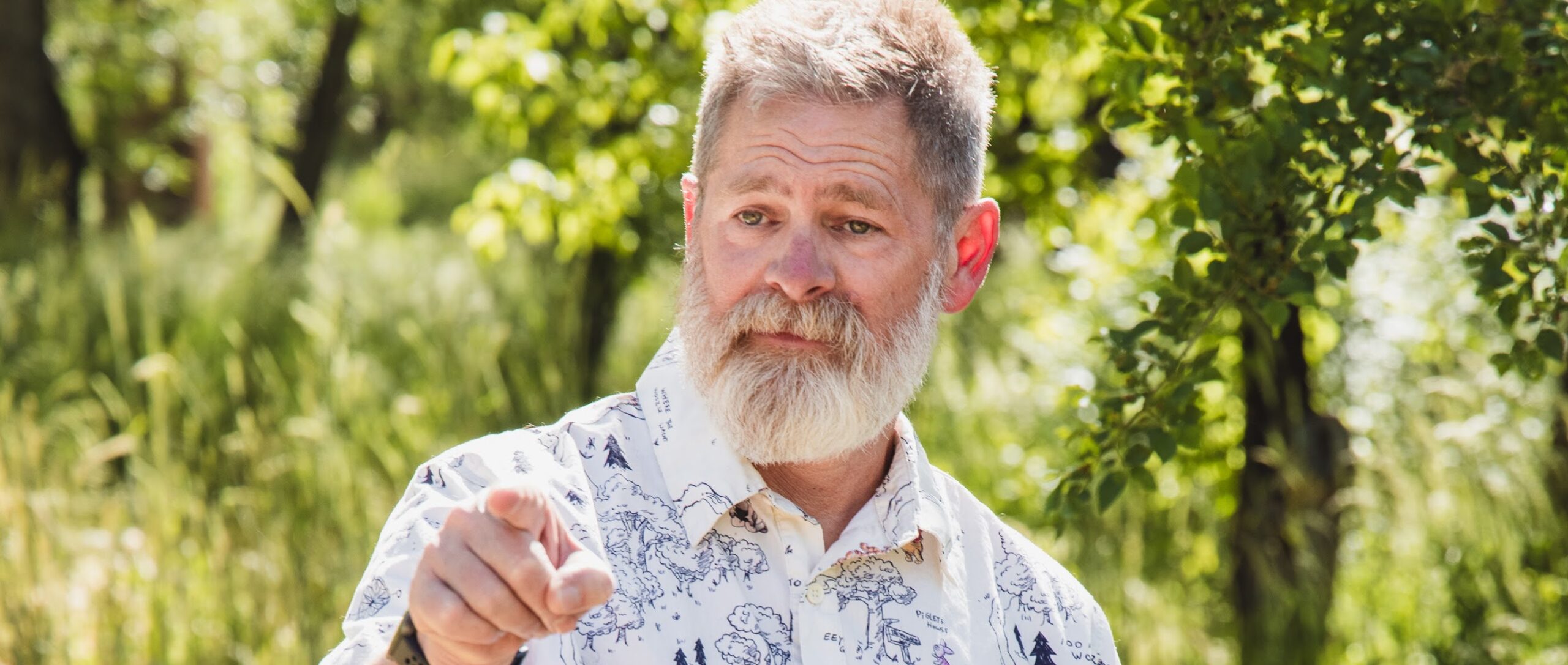 Kevin stands outdoors in a sunlit wooded area, pointing toward something off camera with a thoughtful expression.