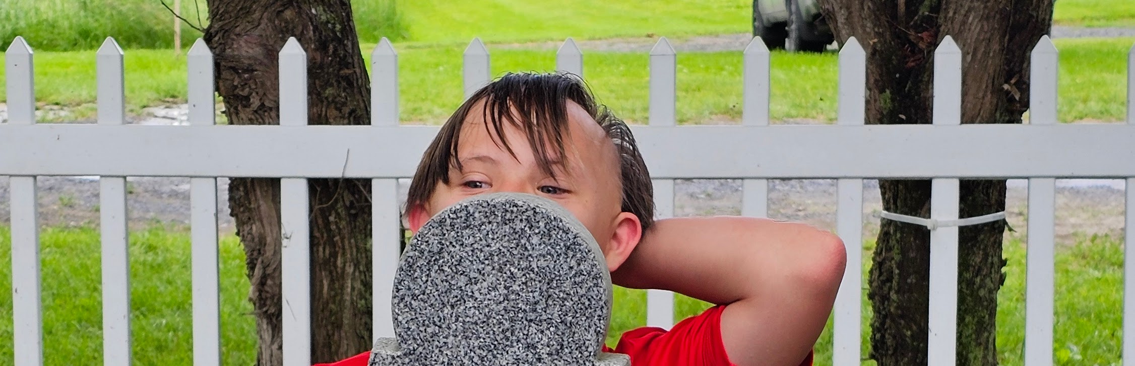 Ben peeks over the top of a gray sign with one arm resting behind his head, framed by a white picket fence and green grass.
