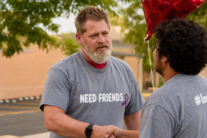 A bearded man wearing a gray “Needs Friends” T-shirt shakes hands with another man outdoors beneath trees, with a red balloon nearby.