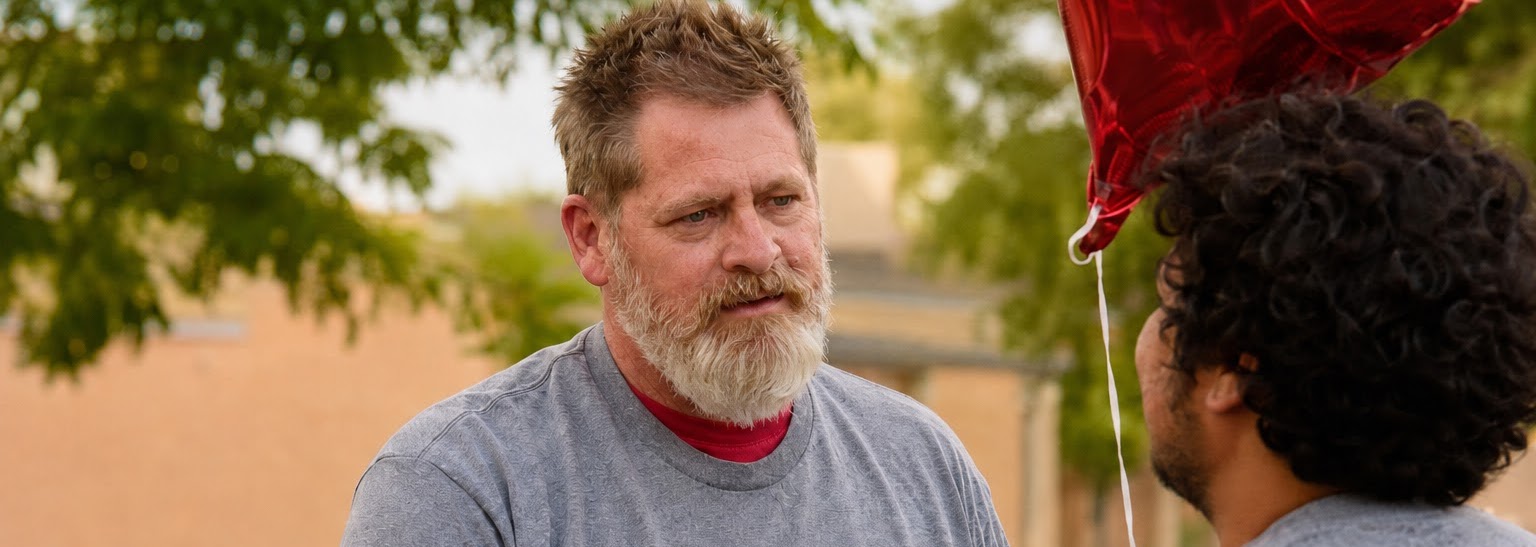 A bearded man wearing a gray “Needs Friends” T-shirt shakes hands with another man outdoors beneath trees, with a red balloon nearby.