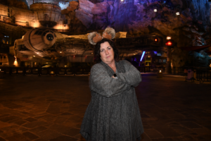 Cindy stands with her arms crossed in front of the Millennium Falcon at a Disney park, wearing themed ears and looking directly at the camera.
