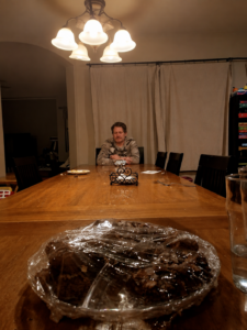 A man sits quietly at the far end of a long family dining table after a meal, surrounded by empty chairs, dishes, crumbs, and everyday signs of a busy home.