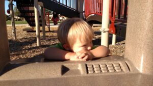 A young boy rests his folded arms on playground equipment, looking quietly to the side as warm afternoon sunlight falls across his face.