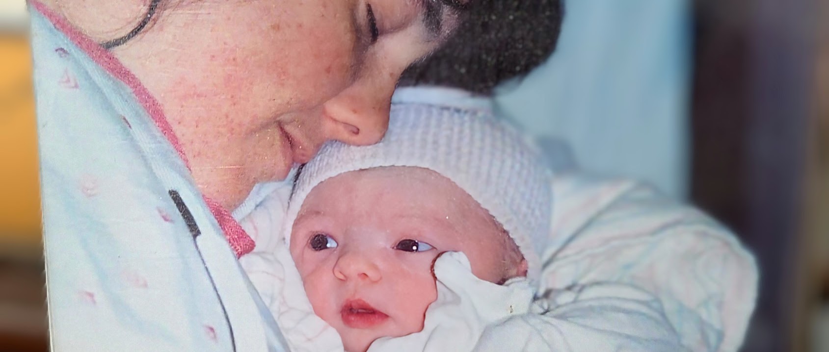 Close-up of a young mother resting her face against her newborn baby, who is wrapped in a blanket and wearing a soft knit cap.
