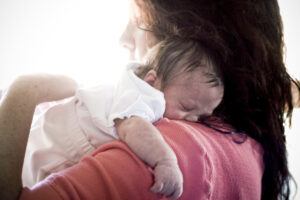 Cindy holds a sleeping newborn against her shoulder, cradling the baby close in soft natural light.