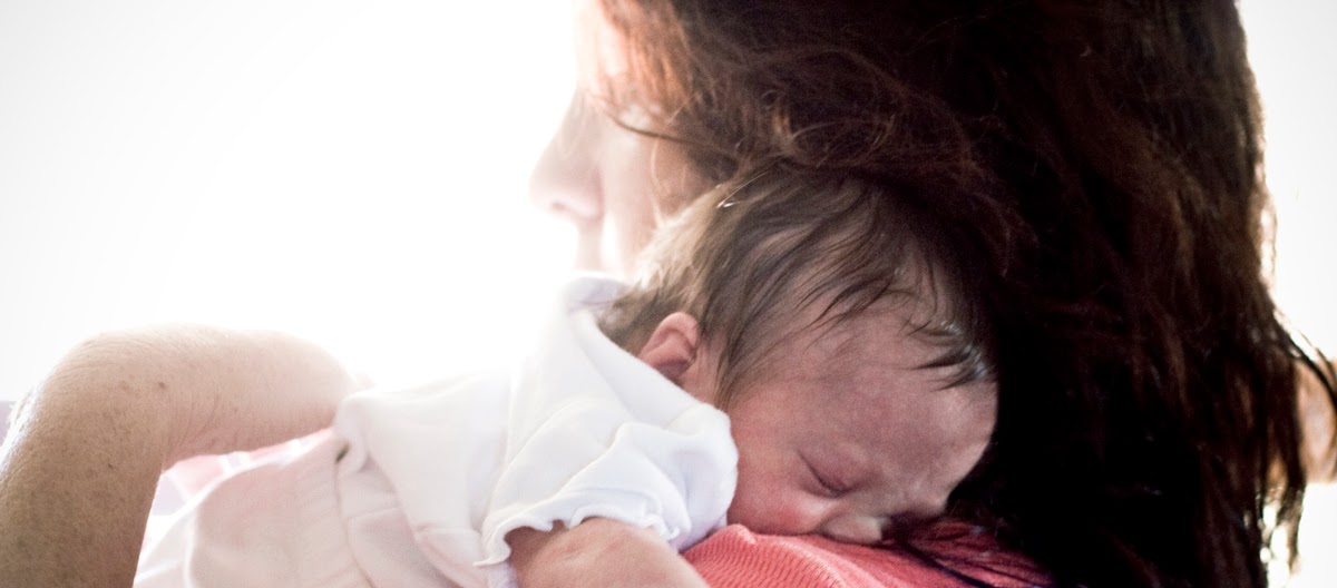 Cindy holds a sleeping newborn against her shoulder, cradling the baby close in soft natural light.